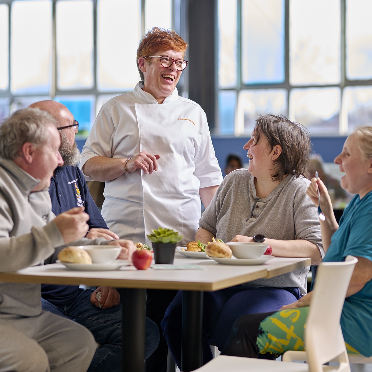 A group of four people with three sat around a table and one standing up wearing an apron