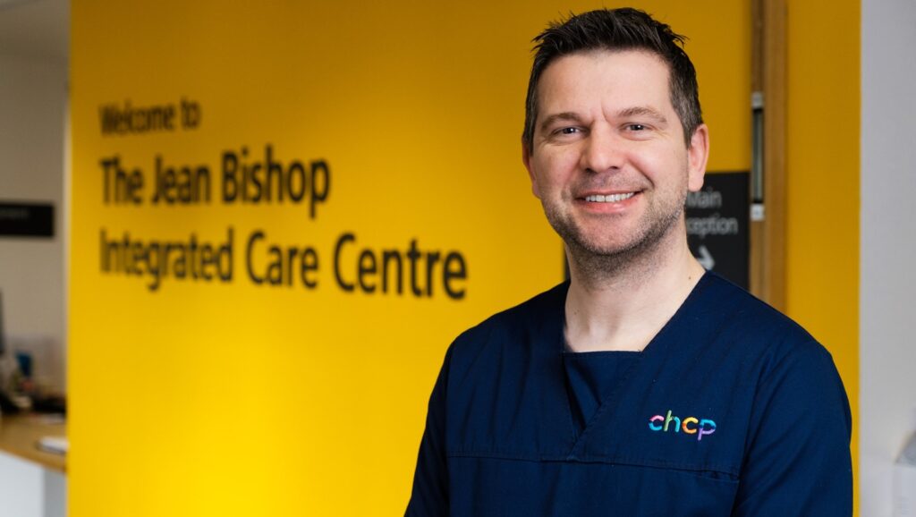A doctor from City Healthcare Partnership in medical overalls. Behind him is a yelllow wall with the wording The Jean Bishop Integrated Care Centre