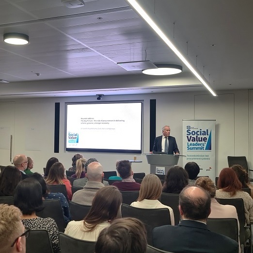A man speaking at a podium with a white banner behind him with the wording Social Value Leaders' Summit in blue
